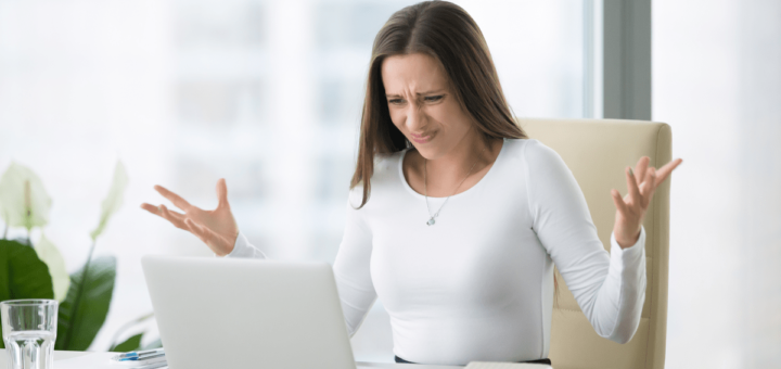 Junge Geschäftsfrau sitzt verärgert am Büroschreibtisch vor Laptop und hebt verzweifelt die Hände.